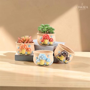 May include: A set of four beige ceramic planters with textured surfaces, each featuring raised, colorful mushroom designs. One planter holds a green succulent, while the others are empty. The brand name OAKIRA goods is printed in the top right corner.