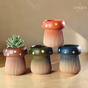 May include: Four mushroom-shaped ceramic planters with ribbed stems and spotted caps in orange, brown, green, and blue. One planter contains a green succulent. The text OAKIRA goods is visible in the upper right corner of the image.