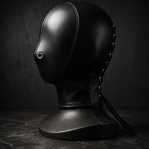 May include: A black leather hood featuring a smooth, rounded design that covers the head and neck. The hood includes a single circular metal eyelet positioned at the nose area and a lace-up closure with metal grommets running down the back.