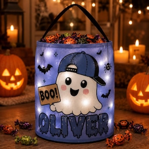 May include: A light blue cylindrical Halloween candy bucket with a black handle, featuring an illuminated ghost wearing a blue baseball cap, holding a sign that says BOO!, and the name OLIVER printed in large dark blue letters at the bottom.