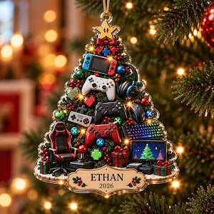 Peut inclure: Une décoration de sapin de Noël triangulaire sur le thème du jeu vidéo avec des manettes miniatures, un casque, un clavier, une chaise de jeu et un écran. L'ornement comporte une plaque en bois à la base avec le texte ETHAN et 2026.