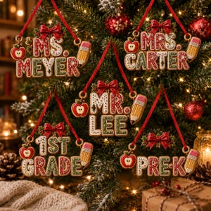 Op de afbeelding: Vijf hangende kerstornamenten met rode en groene letters met witte accenten. Elk ornament toont een naam of leerjaar zoals Ms. Meyer, Mrs. Carter, Mr. Lee, 1st Grade en Pre-K, versierd met een rode strik, een appel en een potlood.