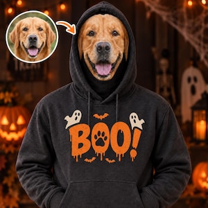 以下が含まれることがあります： オレンジ色で「BOO!」と書かれたグラフィックプリントのダークグレーのパーカー。Oの文字は黒い肉球のマークになっています。デザインには小さな白い幽霊とオレンジ色のコウモリが含まれています。フードの中にゴールデンレトリバーの顔が見えます。