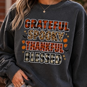 Op de afbeelding: Een antracietgrijs sweatshirt met ronde hals en een grafische print met de woorden GRATEFUL in rood ruitpatroon, SPOOKY in luipaardprint, THANKFUL in oranje en zwart, en BLESSED in beige en zwart ruitpatroon, versierd met kleine pompoenen, vleermuizen en bladeren.