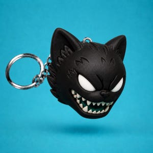 May include: A black cat head keychain featuring a matte finish, large white almond-shaped eyes, and a wide grin displaying sharp white teeth. The keychain includes a silver-toned metal split ring attached by a small chain to the top of the head.
