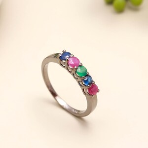 May include: A silver-toned ring featuring five small, round, faceted gemstones set in a row. The stones are arranged in a sequence of blue, pink, green, blue, and pink, held in place by a simple metal band with a polished, reflective finish.