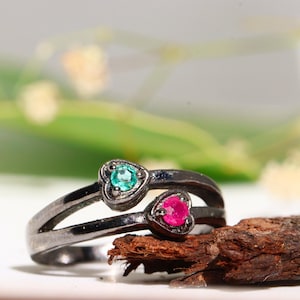 May include: A dark-toned metal ring featuring a split band design. Two heart-shaped settings are positioned at the ends of the split, with one containing a round, vibrant green gemstone and the other containing a round, bright pink gemstone.