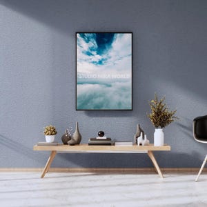 Op de afbeelding: Een rechthoekige wandkunstprint in een zwarte lijst met een blauwe lucht met witte wolken en de tekst STUDIO MIRA WORLD. De print hangt boven een lichthouten bankje met kleine vazen, een plant in een pot en gestapelde boeken.