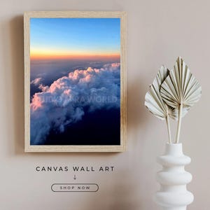 Op de afbeelding: Een rechthoekige ingelijste wandkunstprint met een luchtfoto van wolken bij zonsondergang met een gradiëntlucht die overgaat van oranje naar blauw. De tekst STUDIO MIRA WORLD is zichtbaar op de print. Onder de lijst staat de tekst CANVAS WALL ART, een pijl naar beneden en een knop met SHOP NOW.