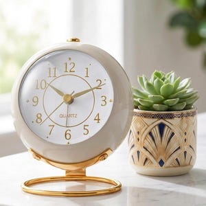 May include: A round cream-colored quartz table clock with gold-tone hands and numerals, mounted on a circular gold metal stand. Beside it sits a small cylindrical planter featuring a cream, gold, and navy blue geometric art deco pattern.