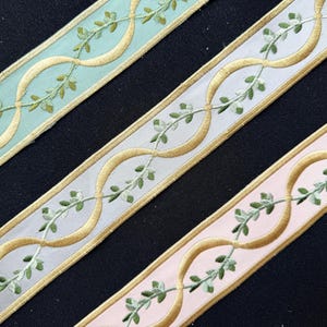 May include: Three strips of decorative ribbon featuring a repeating gold embroidered wave pattern intertwined with a delicate green leaf vine. The ribbons are presented in three distinct base colors: light mint green, pale gray, and soft pastel pink.
