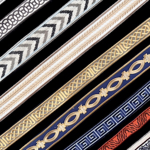 May include: A collection of decorative fabric trim ribbons arranged diagonally. The ribbons feature various patterns including white floral scrolls, black and white chevrons, gold Celtic knots, blue and gold ovals, and red and black wave designs.