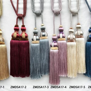 May include: Six decorative curtain tiebacks with tassels, labelled ZMDSA17-1 through ZMDSA17-6. Each features a braided cord, a decorative knot, and a dual-tassel design in gold, burgundy, grey, mauve, cream, and blue with matching cord patterns.
