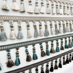 May include: Four horizontal strips of decorative tassel fringe trim. The top row features light cream and pale pink tassels. The second row has silver and grey tassels. The third row displays teal and taupe tassels. The bottom row shows navy and taupe.