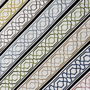 May include: A collection of fabric trim strips arranged diagonally, each featuring a white base with a repeating geometric lattice pattern embroidered in various colors including gold, gray, blue, green, tan, navy, and soft pink.