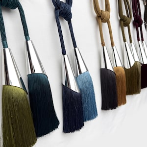 Pode incluir: Uma coleção de braçadeiras para cortinas com cordões trançados e ponteiras cónicas de metal prateado. As borlas estão disponíveis em várias cores, incluindo verde-azeitona, azul-escuro, azul-marinho, azul-claro, bege, castanho e bordeaux.