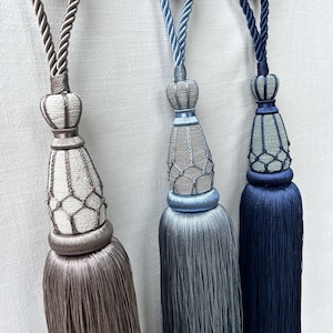 May include: Three decorative curtain tiebacks featuring long, silky tassels in shades of taupe, light blue, and navy blue. Each tieback includes a braided cord loop, a decorative knot, and a woven net-like structure covering the upper tassel section.