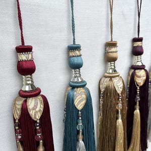 Op de afbeelding: Een rij decoratieve gordijnkwasten in diverse kleuren, waaronder bordeauxrood, teal, goud, donkerpaars, zilver en bruin. Elke kwast is voorzien van een gevlochten ophangkoord, een cilindrische bovenkant met strass-accenten en fijne draden.
