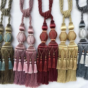 May include: Five decorative curtain tiebacks featuring twisted rope cords and dual tassels. Each tieback includes a central bulbous ornament wrapped in fine thread netting, finished with smaller hanging fringe tassels in olive, pink, red, gold, and gray.