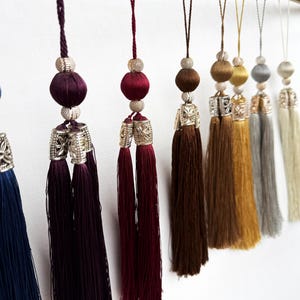 May include: A collection of decorative hanging tassels in various colors including deep blue, purple, burgundy, brown, gold, silver, and cream. Each tassel features a braided hanging cord, a round bead, and a metallic cap with floral detailing.