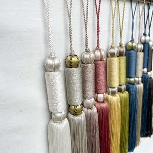 May include: A collection of decorative fabric tassels hanging from a gold-toned metal rod. The tassels feature various colors including white, silver, beige, rose, gold, yellow, blue, and brown, each with a looped cord and cylindrical top.