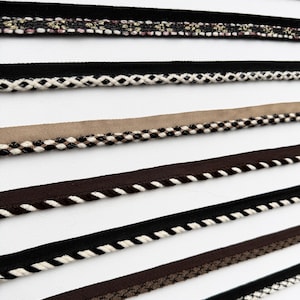 May include: A collection of decorative trim ribbons arranged in parallel rows. The assortment includes various textures and patterns, featuring black, brown, beige, and metallic silver threads woven into intricate braided and geometric designs.
