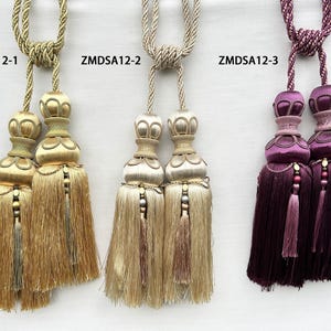 May include: Three decorative curtain tiebacks with tassels, labeled ZMDSA12-1, ZMDSA12-2, and ZMDSA12-3. The first is gold, the second is champagne beige, and the third is deep purple. Each features a braided cord, a knotted top, and layered fringe.