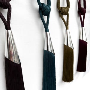 May include: Four decorative curtain tiebacks featuring knotted rope cords in dark brown, teal, olive green, and burgundy. Each tieback includes a polished silver-toned metal cone cap that secures a long, straight fringe tassel in a matching colour.