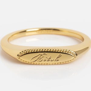 May include: A gold-toned ring featuring an oval-shaped face with a beaded border. The center of the oval is engraved with the word "Bitch" in a cursive script font. The band is smooth and tapers toward the back of the ring.