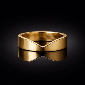 Könnte beinhalten: Ein polierter Goldring mit einer ausgeprägten V-förmigen Vertiefung an der Oberkante. Der Ring hat eine glatte, reflektierende Oberfläche und eine solide Konstruktion, die ein durchgehendes metallisches Finish zeigt, das das Licht gleichmäßig reflektiert.