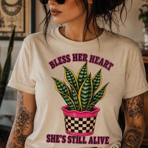 Peut inclure: Un t-shirt beige à manches courtes avec un motif représentant une plante serpent verte dans un pot à carreaux roses et noirs. Le texte "BLESS HER HEART" est imprimé en rose au-dessus de la plante et "SHE'S STILL ALIVE" en dessous du pot.
