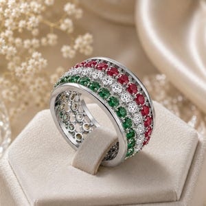 Puede incluir: Un anillo ancho de tono plateado con tres filas paralelas de piedras de corte redondo. Las filas consisten en gemas de color rojo vibrante, blanco transparente y verde intenso engastadas en la superficie de metal pulido, creando un efecto de rayas multicolor.