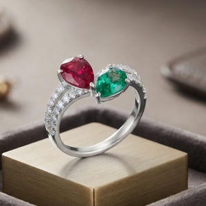 Può includere: Un anello in argento con due pietre preziose a taglio a goccia, una rosso intenso e una verde brillante, posizionate fianco a fianco. Il gambo è diviso e decorato con piccole pietre bianche rotonde lungo le spalle, creando un design bypass.