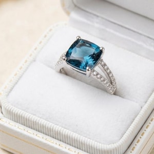 May include: A silver ring featuring a large, cushion-cut, deep blue gemstone set in a four-prong mount. The band is split into two sections, each encrusted with small, clear, round-cut stones that extend along the sides of the ring shank.