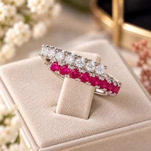 May include: A bypass ring featuring two parallel bands of round stones. One band consists of clear, colourless stones, while the other band features vibrant, deep pink stones. The metal setting is silver-toned, and the ring is displayed on a beige cushion.