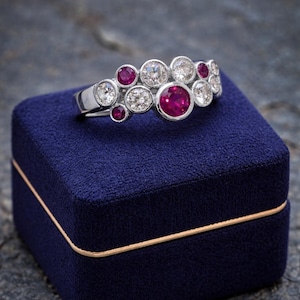 May include: A silver-toned ring featuring a cluster of round-cut stones in a bezel setting. The design includes several clear white diamonds and vibrant red rubies arranged in a staggered, multi-sized pattern across the top of the band.