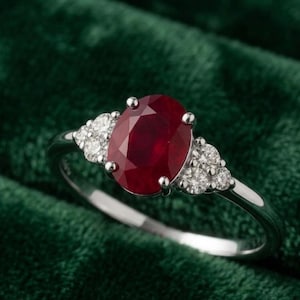 Può includere: Un anello in argento con un grande rubino rosso scuro a taglio ovale come pietra centrale. Il rubino è affiancato su ciascun lato da un gruppo di tre piccoli diamanti bianchi a taglio rotondo incastonati in una fascia d'argento lucida.