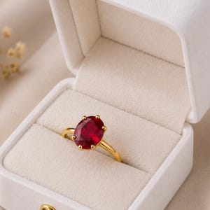 Può includere: Un anello in oro con una grande pietra preziosa rosso intenso dal taglio ovale, fissata da sei griffe. L'anello è esposto all'interno di un portagioie bianco aperto, foderato in velluto, con finitura esterna opaca e una chiusura dorata.