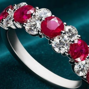 Può includere: Un anello color argento con una fila di rubini rosso vivo a taglio rotondo alternati a diamanti bianchi trasparenti. Le pietre preziose sono incastonate in una fascia di metallo lucido, creando una disposizione scintillante sul gioiello.