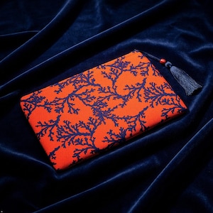 Può includere: Una pochette rettangolare in tessuto con base color arancione vivace e un motivo a corallo ramificato blu scuro. La chiusura a zip è arricchita da una nappa blu scuro e da una piccola perla in legno di colore bruno-rossastro.