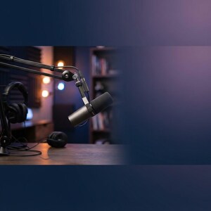 May include: A dark gray professional dynamic microphone mounted on a black adjustable boom arm, positioned above a wooden surface. A pair of black over-ear headphones with a connecting cable rests on the same surface near the microphone base.