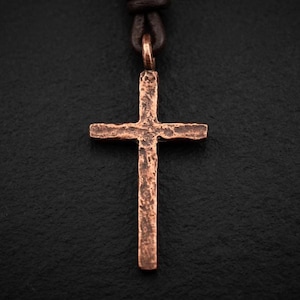 May include: A copper cross pendant with a textured, hammered finish suspended from a dark brown leather cord. The cross features a vertical bar longer than the horizontal bar, showcasing a rustic, uneven surface with deep metallic orange tones.