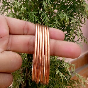 Op de afbeelding: Een set van vijf dunne, gepolijste koperen armbanden met een gladde, reflecterende metalen afwerking. De armbanden zijn cirkelvormig en hebben een consistent, eenvoudig ontwerp zonder extra patronen, gravures of decoratieve elementen.
