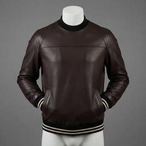 Puede incluir: Una chaqueta tipo suéter de cuero marrón oscuro con cuello redondo acanalado negro, mangas largas y puños y dobladillo acanalados negros con rayas blancas finas. La prenda incluye dos bolsillos laterales y una costura horizontal en el pecho.