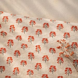 May include: A light beige fabric featuring a repeating pattern of small, stylised orange trees with dark brown trunks and branches. A small, dried orange-coloured floral sprig rests on the lower right side of the textured textile surface.