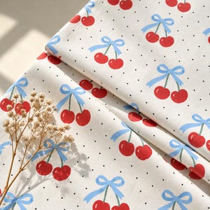 May include: A white fabric featuring a repeating pattern of red cherries paired with light blue bows and small black polka dots. The textile is folded, displaying the cherry and bow print across the surface of the material.