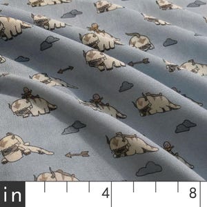 May include: Light blue fabric featuring a repeating pattern of cream-colored flying bison creatures with brown arrows and small figures riding on their backs, interspersed with dark gray clouds and brown arrows, shown with a ruler measuring 8 inches.