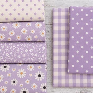 Peut inclure: Une collection de six pièces de tissu pliées dans des tons lavande et blanc. Les motifs incluent des pois violets sur fond crème, des marguerites blanches sur lavande, des imprimés floraux blancs, des marguerites à cœur noir et un vichy lavande.