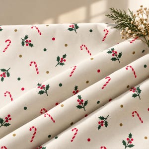 May include: A cream-coloured fabric featuring a repeating holiday pattern of red and white striped candy canes, green holly leaves with red berries, and small scattered polka dots in shades of dark green and metallic gold across the surface.