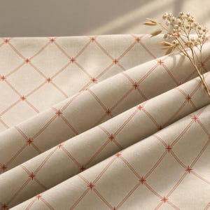 Peut inclure: Un morceau de tissu écru plié présentant un motif répétitif de fines lignes orange entrecroisées formant un quadrillage en losanges, avec de petites formes d'étoiles orange à chaque intersection, accompagné d'un brin de fleurs blanches séchées.